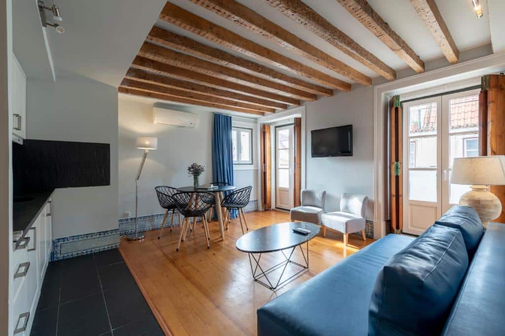Sala de estar do Living Lisboa Baixa Apartments com vigas de madeira, sofá azul, mesa de jantar redonda com cadeiras, TV na parede e janelas com cortinas azuis.