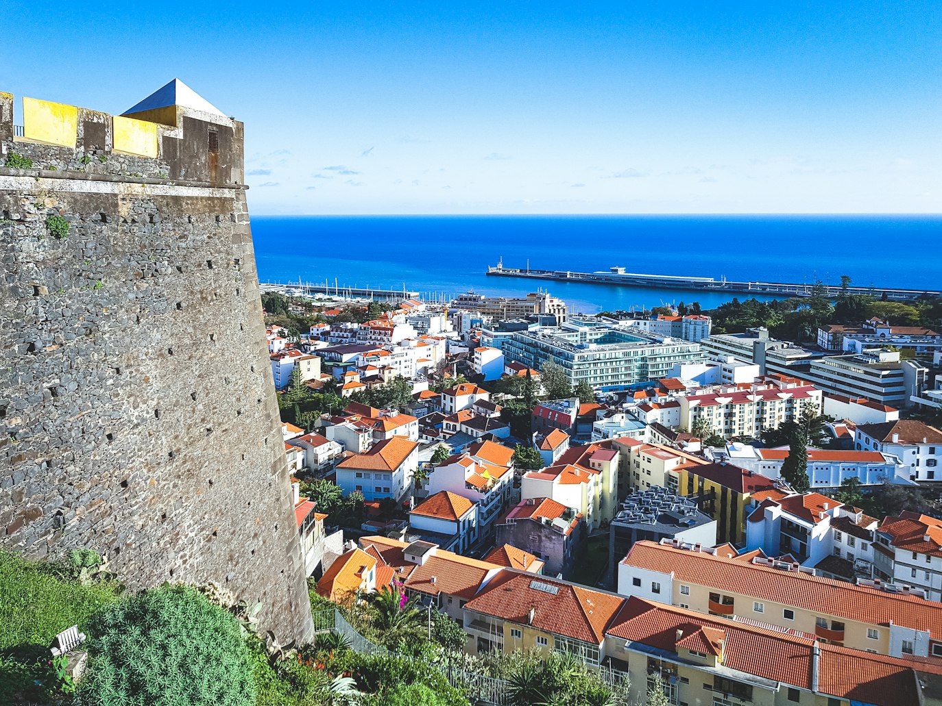 Vista elevada de uma paisagem urbana costeira, com edifícios coloridos, um muro de pedra e um porto.