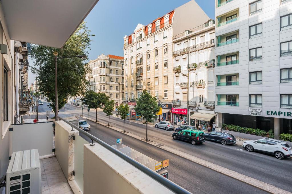 Vista de uma varanda do 137 LuxResidence sobre uma rua da cidade com carros estacionados, árvores e prédios de vários andares sob um céu azul claro.