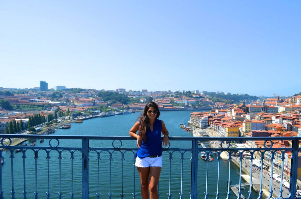 Uma pessoa no parapeito de uma ponte no Porto, com vista para o rio e para a paisagem urbana sob um céu azul.