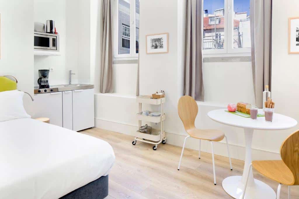 Quarto iluminado do Ascensor da Bica - Lisbon Serviced Apartments com cama, cozinha compacta e zona de refeições. Inclui mesa pequena, duas cadeiras, um carrinho e duas janelas com cortinas.
