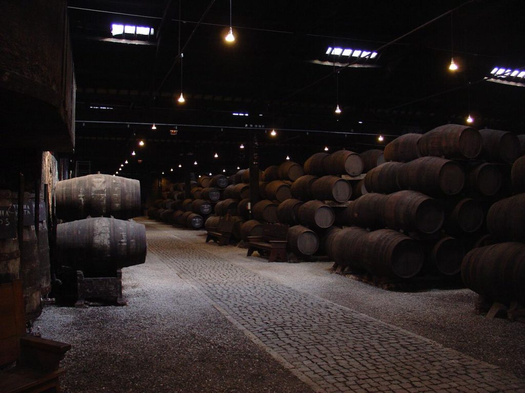 Cave de Vinho do Porto pouco iluminada, com fileiras de grandes barris de madeira ao longo de um caminho de calçada.