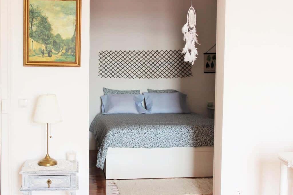 Quarto acolhedor da Casa da Varanda Simpática com cama coberta por roupa de cama estampada em azul, um apanhador de sonhos, um candeeiro, um quadro emoldurado e um pequeno tapete.