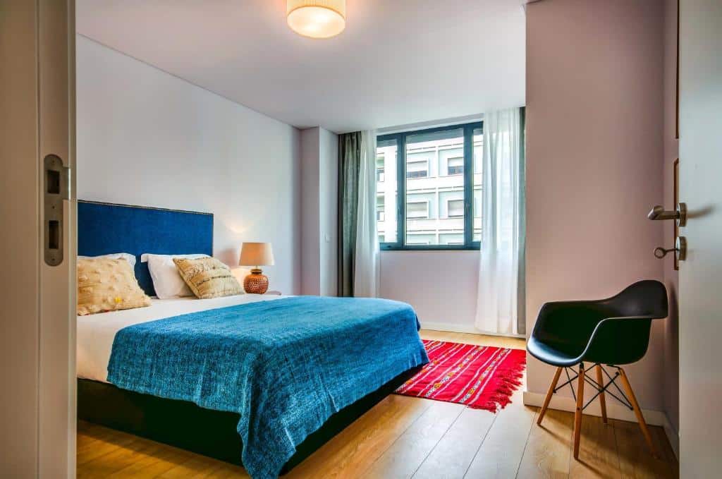 Quarto do Lisbon Serviced Apartments com cama, manta azul e almofadas. Há um candeeiro, uma janela com cortinas e uma cadeira junto à porta.