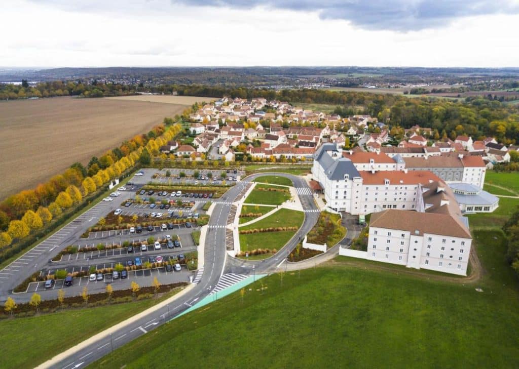 Vista aérea de um grande complexo de edifícios com estacionamentos, rodeado por campos e por uma zona residencial. Este é o B&B Hôtel, um dos hotéis perto da Disney Paris.