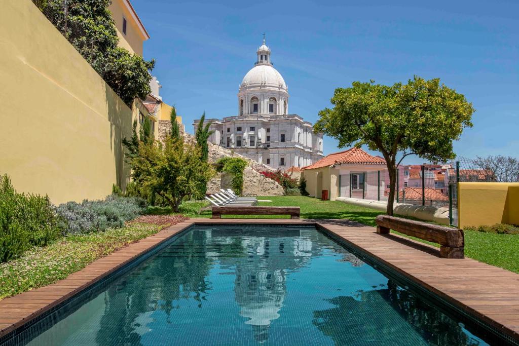 Piscina estreita do Tandem Palacio Alfama Suites rodeada de vegetação e um banco, com um edifício de cúpula ao fundo, sob um céu azul claro.