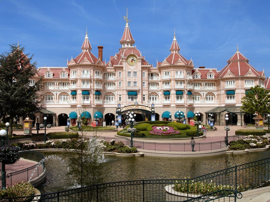 Grande hotel cor-de-rosa com torre de relógio e telhados de mansarda, em frente a uma fonte e a um jardim de flores, sob um céu azul. Este é o Disneyland Hotel, um dos hotéis perto da Disney Paris.