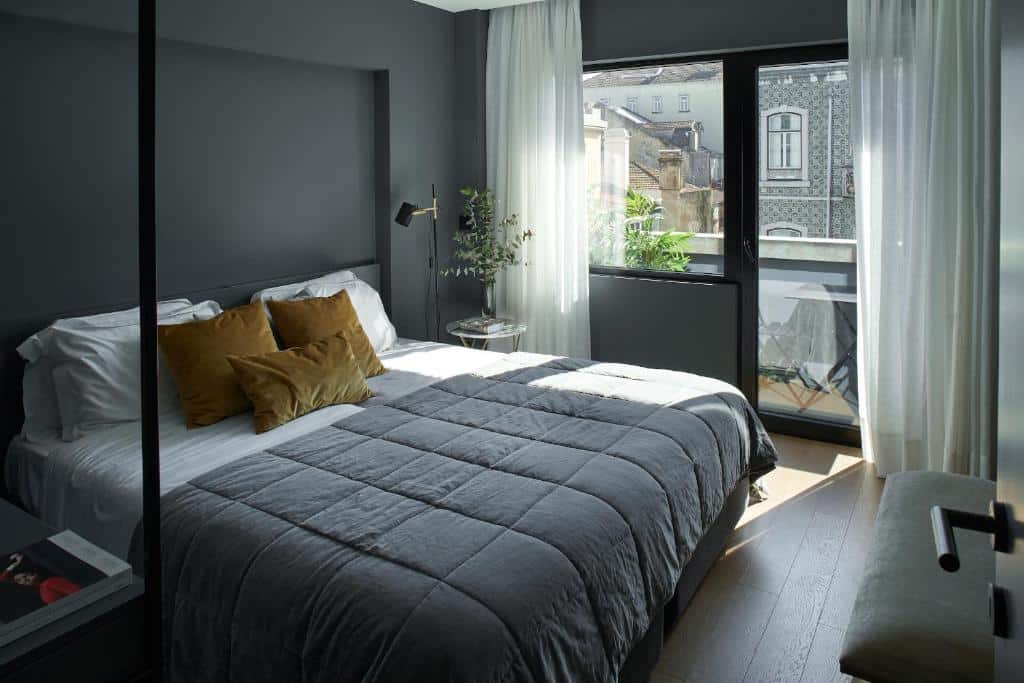 Quarto do Nôma Living Pop Saldanha com cama bem feita, roupa de cama cinzenta e almofadas douradas. Uma grande janela com cortinas transparentes deixa entrar luz natural.