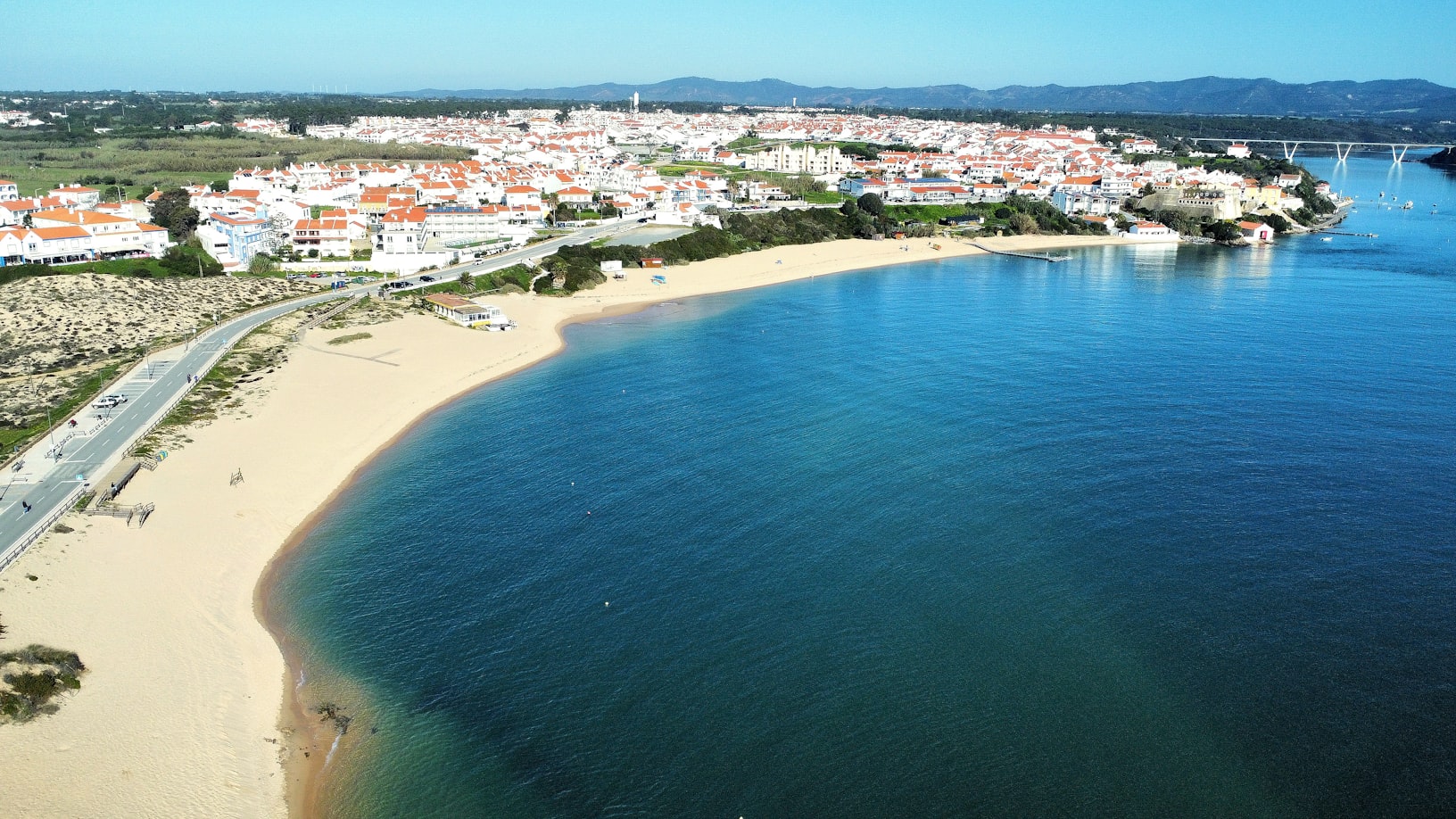 Vista aérea de Vila Nova de Milfontes, com edifícios brancos e telhados vermelhos, praia de areia dourada e mar azul.