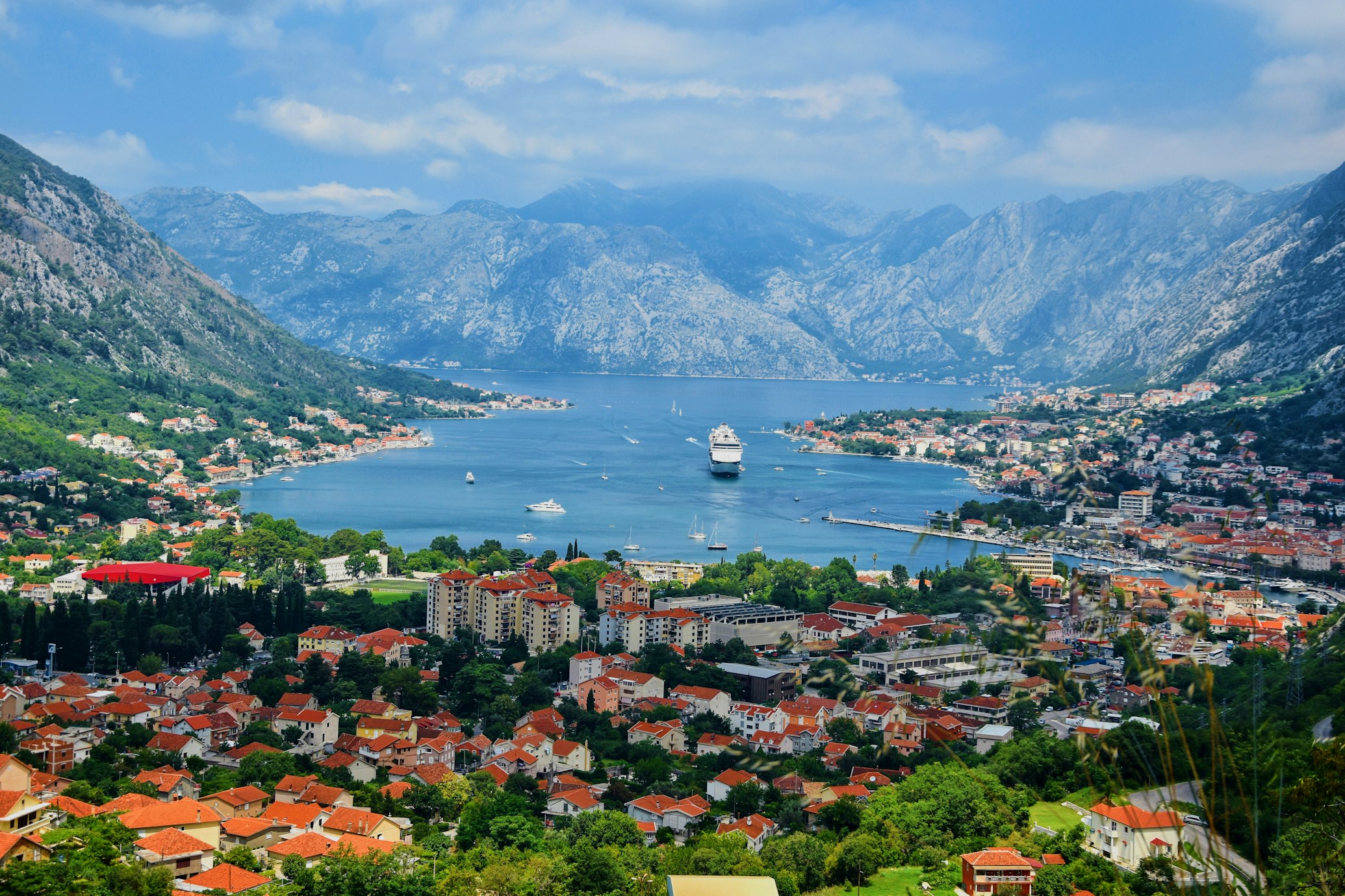 Vista panorâmica da Baía de Kotor, no Montenegro, com edifícios de telhados vermelhos junto à costa, um navio de cruzeiro ao longe e montanhas sob um céu parcialmente nublado.