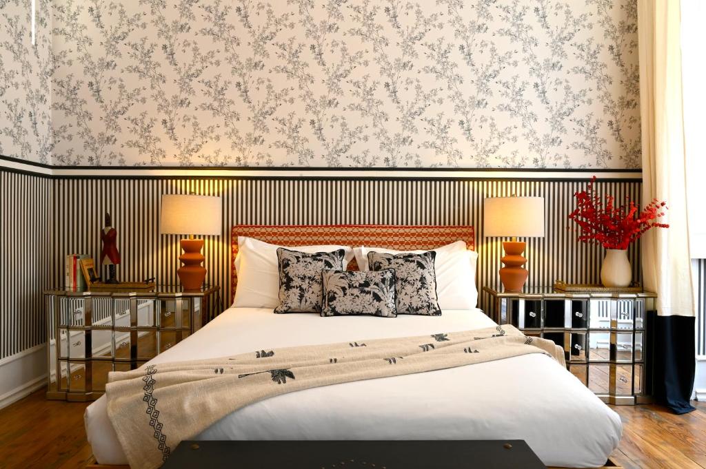 Quarto do Sapateiros 44 com cama, almofadas florais e mesas de cabeceira. O papel de parede tem um padrão floral e listrado. Candeeiros e flores vermelhas completam a decoração.