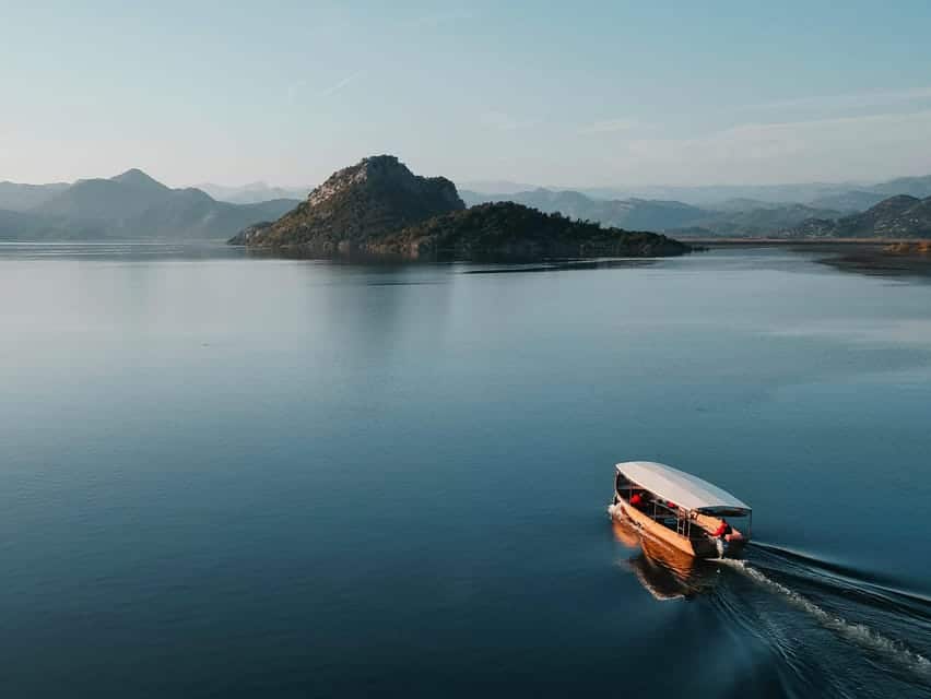 Um barco com cobertura navega no Lago Skadar, no Montenegro, em direção a uma pequena ilha com colinas ao longe, sob um céu limpo.