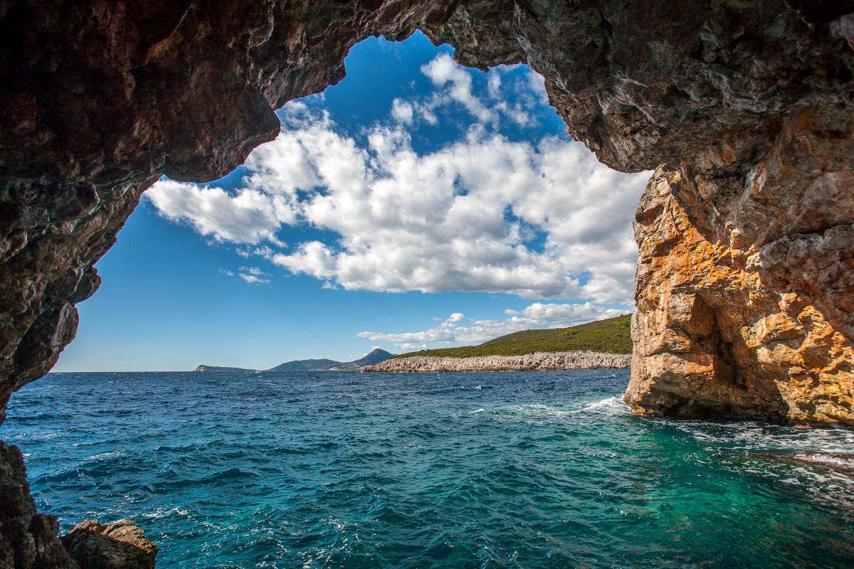 Vista do interior de uma gruta rochosa, conhecida como Gruta Azul, em Perast, com vista para um mar azul e colinas ao longe sob um céu com nuvens dispersas.