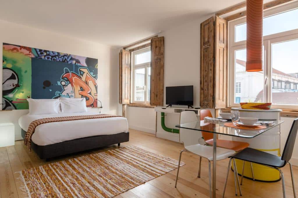 Quarto moderno do Chiado Arty Flats com cama, arte de grafiti na parede, uma pequena mesa de refeições com cadeiras e uma janela com portadas de madeira.