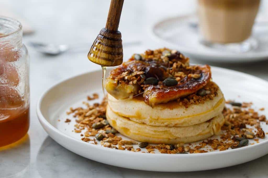 Prato do OnJ S. Lázaro Lisboa com panquecas cobertas de banana caramelizada, nozes, sementes e granola, com mel a ser deitado por uma colher de madeira.