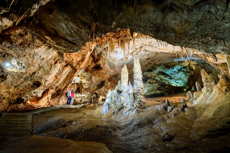 Grande interior da Gruta de Lipa, em Cetinje, com estalactites e estalagmites. Duas pessoas estão à esquerda, admirando as formações rochosas.
