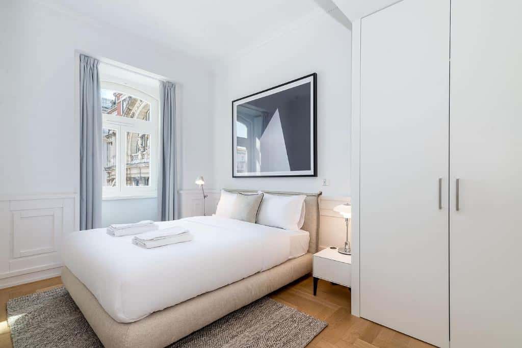 Quarto minimalista do The Boulevard by The Good Neighbour in Lisbon com cama grande, roupa de cama branca, duas toalhas, mesa de cabeceira com candeeiro e um grande roupeiro.