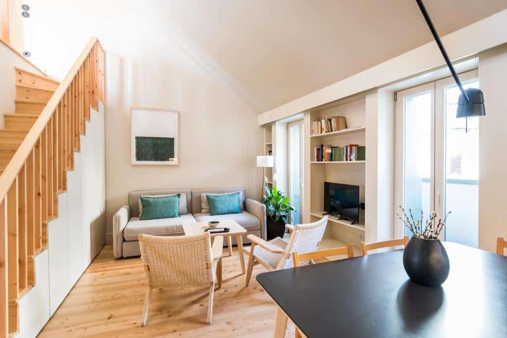 Sala de estar acolhedora das Casas na Portuguesa com sofá, poltronas, uma pequena TV, estantes e uma escada de madeira. Muita luz natural preenche o espaço.