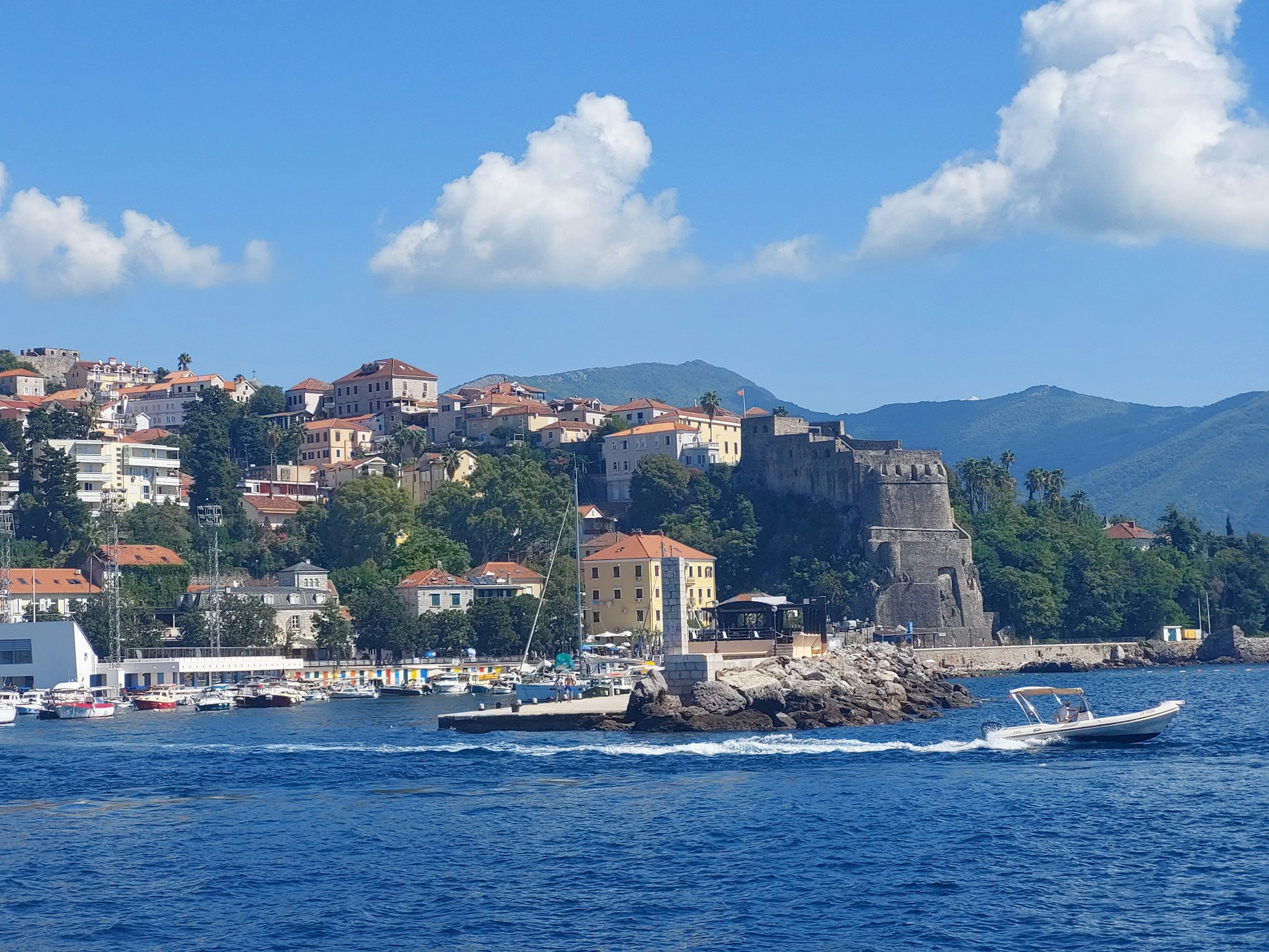 Um pequeno barco navega rapidamente por um mar azul vibrante na cidade costeira de Herceg Novi, com edifícios coloridos e uma fortaleza histórica de pedra.