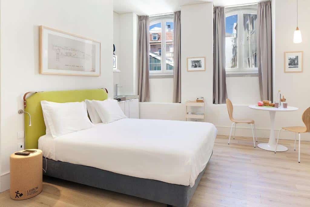 Quarto do Ascensor da Bica - Lisbon Serviced Apartments com cama de casal, mesa pequena e cadeiras. Tem chão de madeira, paredes brancas e duas janelas com cortinas.