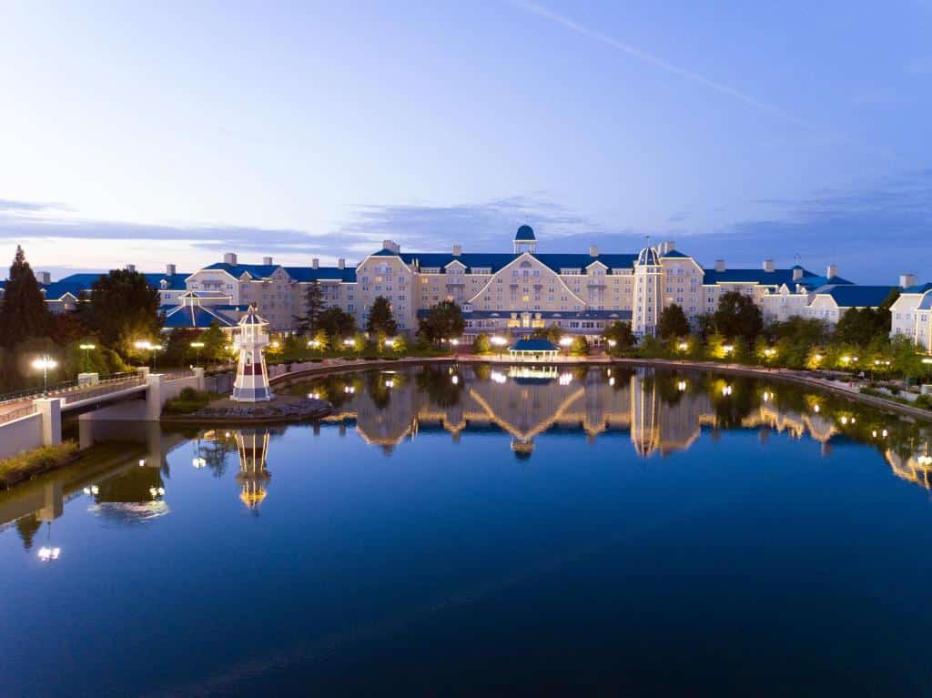 Grande edifício do Disney's Newport Bay Club à beira de água, com telhados azuis, iluminado por luzes exteriores e refletido num lago calmo ao anoitecer.