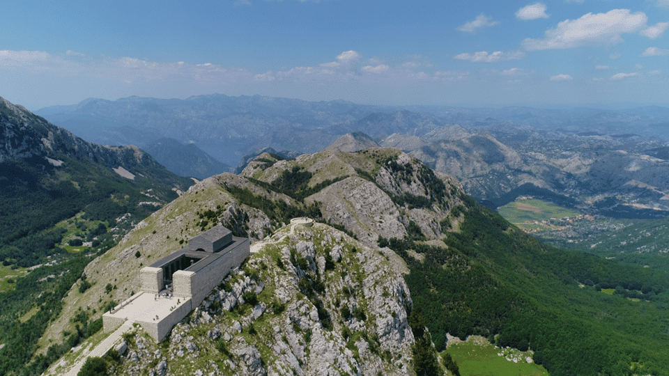 Vista aérea de um edifício de pedra, o Mausoléu de Njegoš, no topo do Monte Lovćen, numa montanha rochosa com vastos vales verdes e cadeias de montanhas ao longe sob um céu azul.