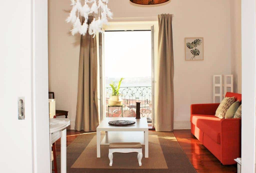 Sala de estar da Casa da Varanda Simpática com sofá cor de laranja, mesa de centro branca e candeeiro de penas. Vê-se uma varanda através de cortinas abertas.