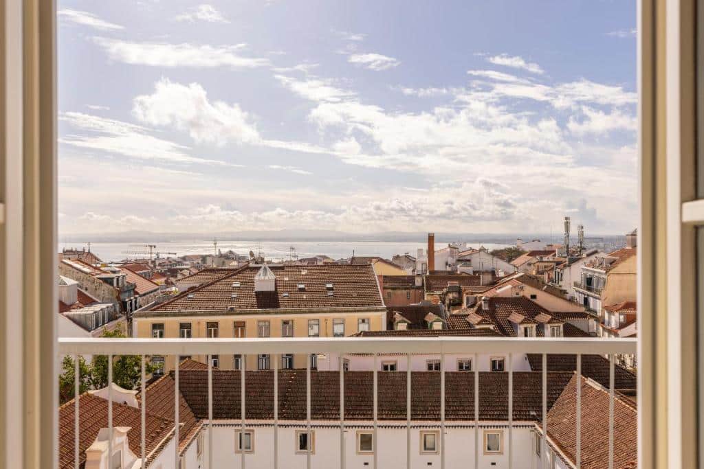 Vista da varanda do Líbere Lisbon Príncipe Real sobre uma paisagem urbana junto à costa, com prédios de telhados vermelhos, céu azul e nuvens.