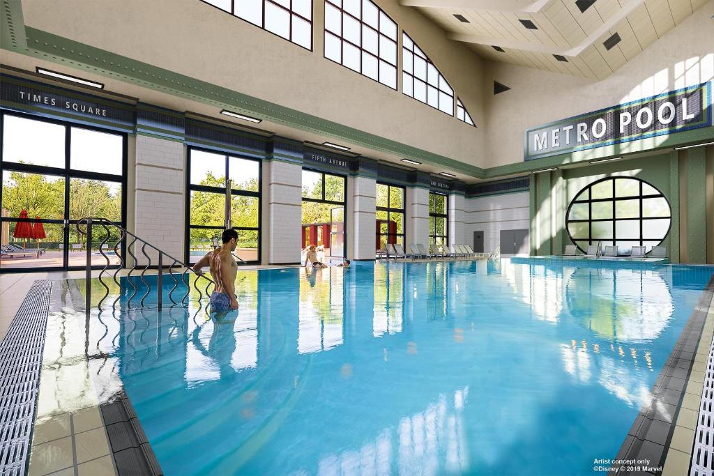Piscina interior do Disney's Hotel New York – The Art of Marvel, com janelas grandes, clarabóias e uma pessoa a entrar na água.