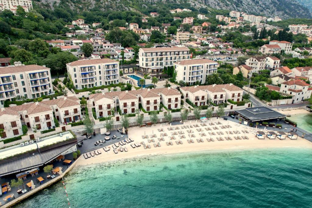 Vista aérea do HUMA Kotor Bay Hotel and Villas, com praia de areia branca repleta de espreguiçadeiras e chapéus de sol e edifícios rodeados por colinas verdejantes.