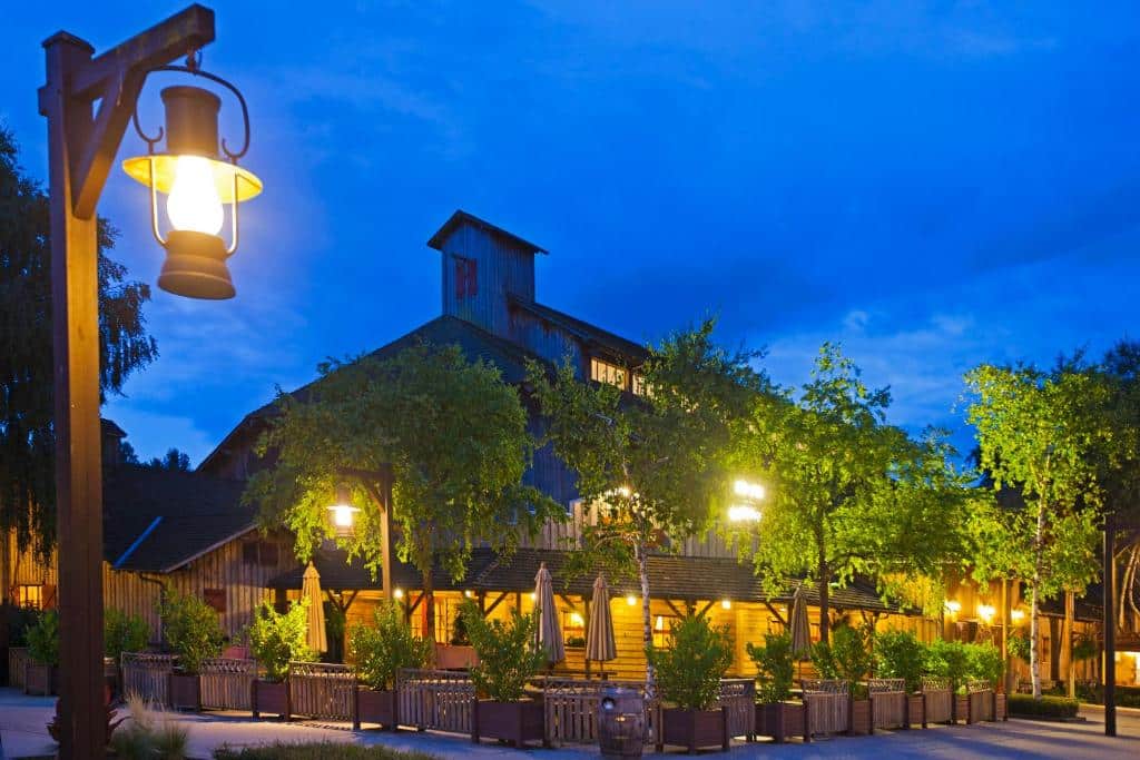 Um edifício rústico em estilo de celeiro com lanternas acesas e árvores em redor, fotografado ao anoitecer com um céu azul profundo. Este é o Disney's Davy Crockett Ranch, um dos hotéis perto da Disney Paris.