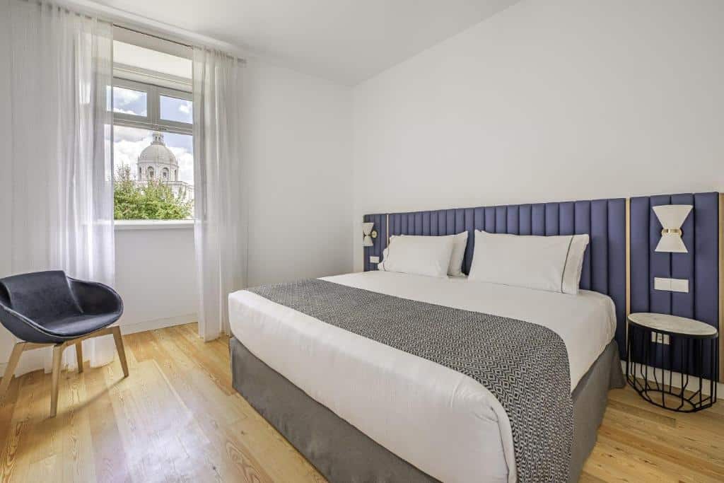 Quarto moderno do Tandem Palacio Alfama Suites com cama grande, cabeceira azul e uma cadeira cinzenta. Uma janela mostra a vista de um edifício com cúpula.