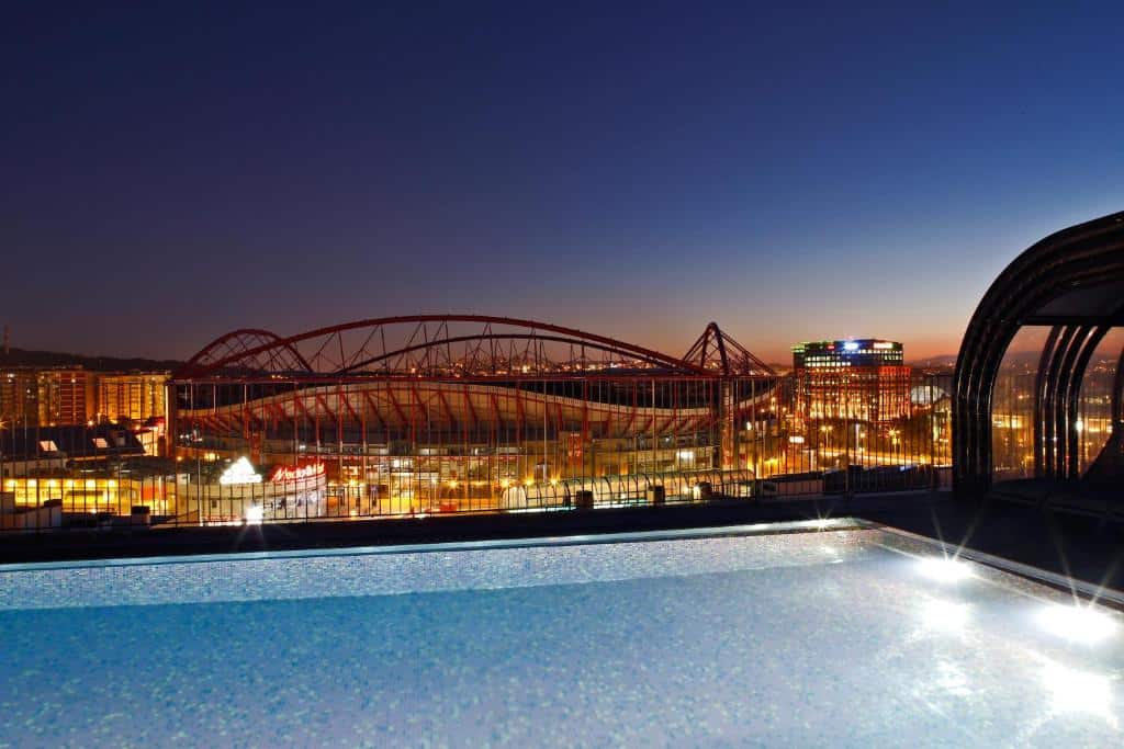 Vista de um estádio iluminado com telhado vermelho à noite, a partir de uma piscina num terraço próximo. As luzes da cidade veem-se ao fundo, no Upon Lisbon Prime Residences.
