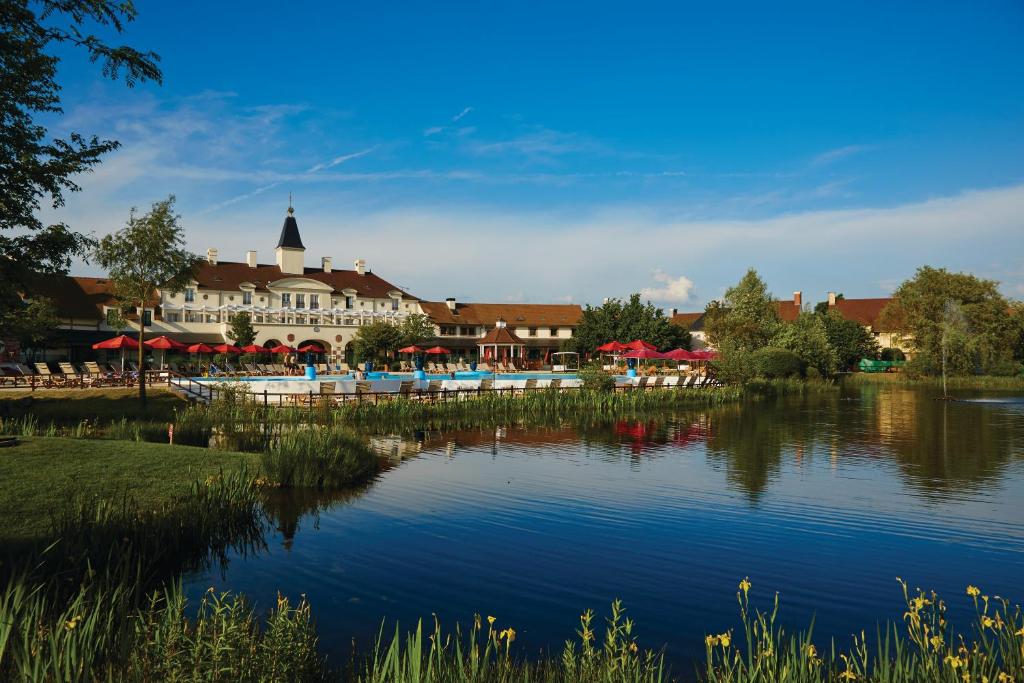 Um grande edifício com chapéus-de-sol vermelhos junto a uma piscina exterior, com vista para um lago rodeado de árvores e relva, sob um céu azul. Este é o Marriott's Village d'Île-de-France, um dos hotéis perto da Disney Paris.