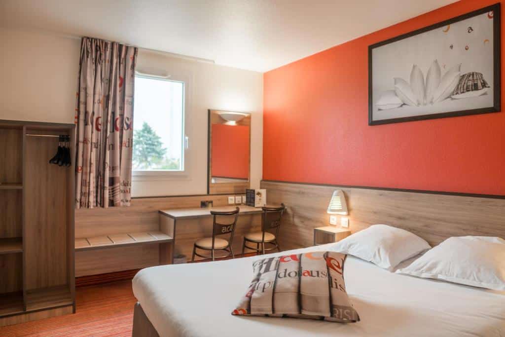 Quarto do Ace Hôtel Paris Marne La Vallée com uma cama de casal, secretária, duas cadeiras, roupeiro aberto, janela e parede laranja com quadros emoldurados.