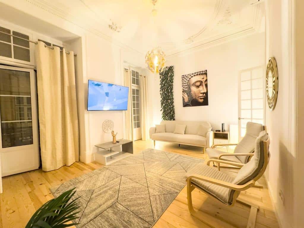 Sala de estar iluminada do Serenity Lisbon Suites com sofá branco, duas cadeiras, TV na parede, uma obra de arte de Buda e grandes janelas com cortinas bege.