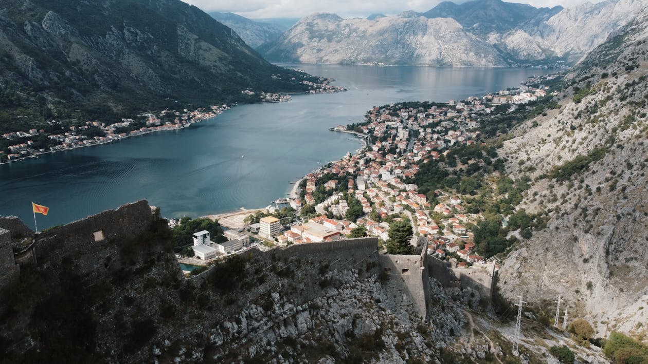 Vista aérea da cidade costeira de Kotor, no Montenegro, com edifícios de telhados vermelhos, rodeada por montanhas e por um grande plano de água, sob um céu nublado.