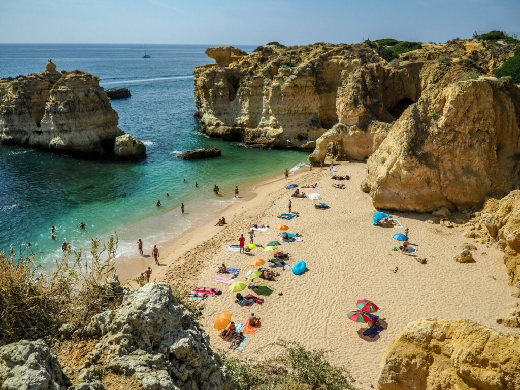 Vista aérea de uma praia no Algarve, rodeada por grandes rochas, com banhistas na areia e mar cristalino.