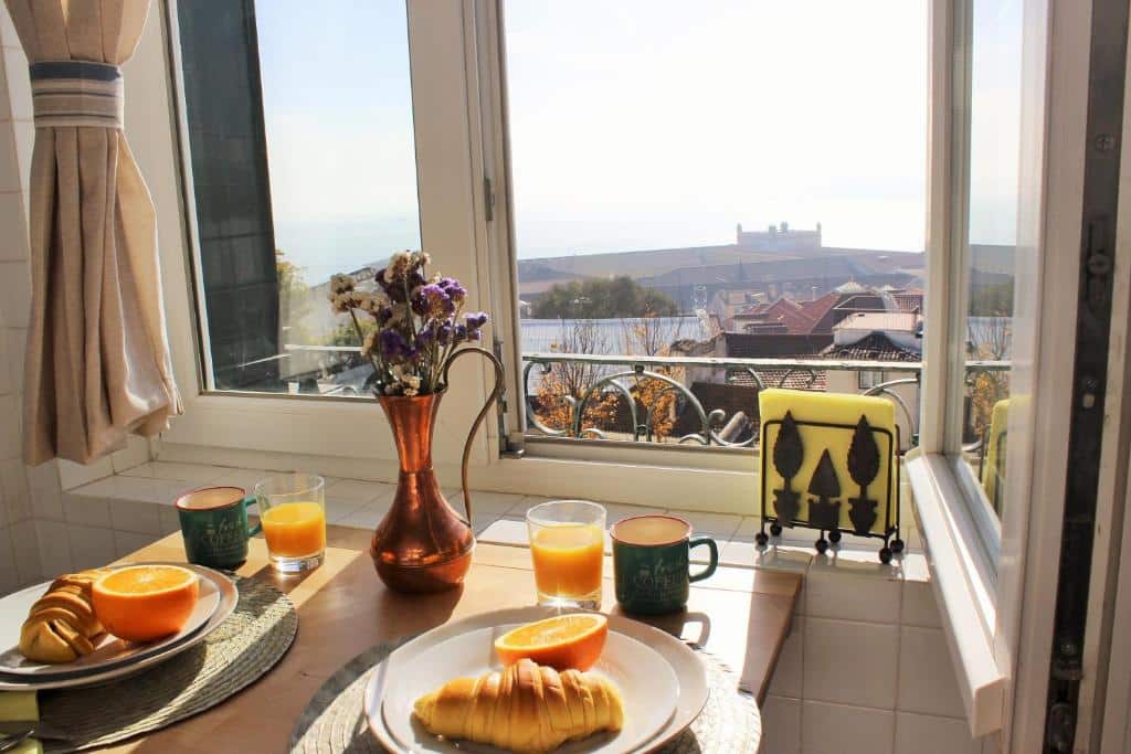 Dois pratos com croissants e dois copos de sumo de laranja numa mesa junto a uma janela, com vista para uma paisagem pitoresca com edifícios, na Casa Rio da Saudade.