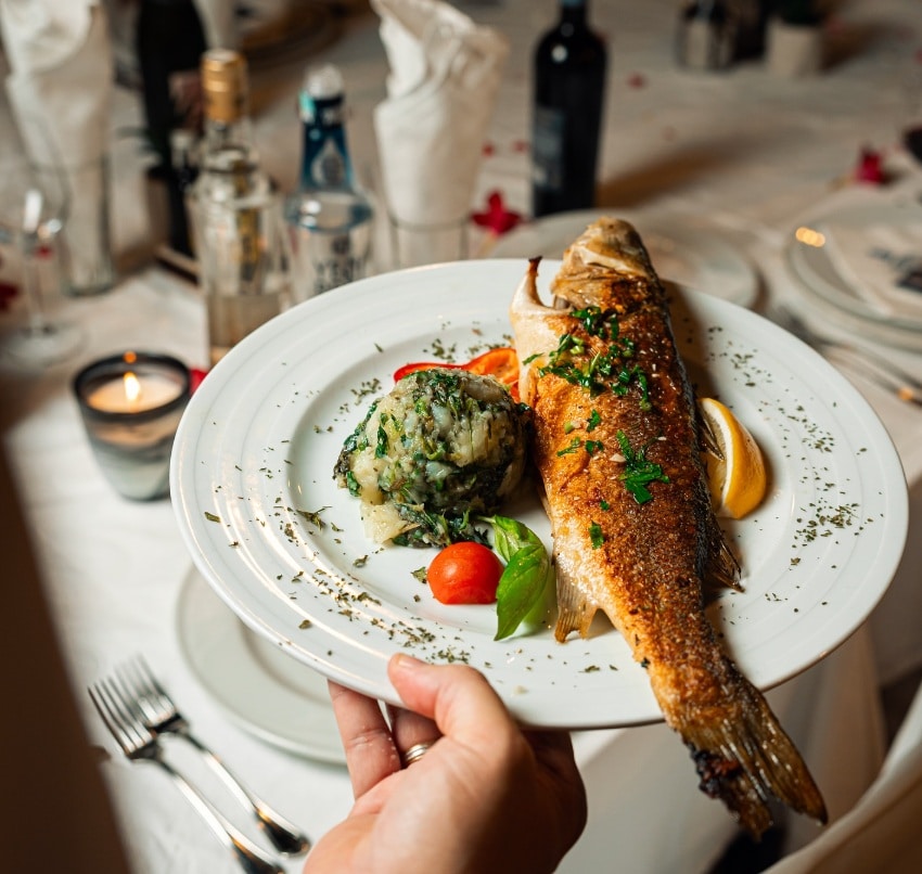 Prato com um peixe inteiro grelhado guarnecido com ervas, uma quenelle de puré de batata com folhas verdes, um tomate-cereja e uma rodela de limão, do restaurante Jadran, em Budva, no Montenegro.
