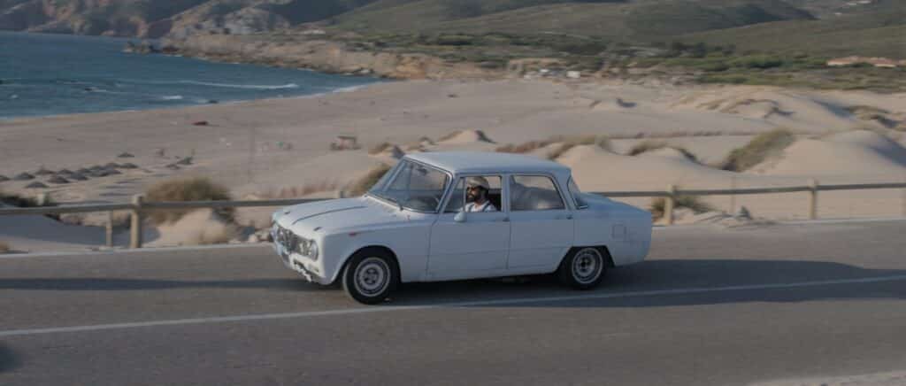 Um automóvel branco antigo percorre uma estrada costeira, com uma praia de areia e falésias rochosas ao fundo.