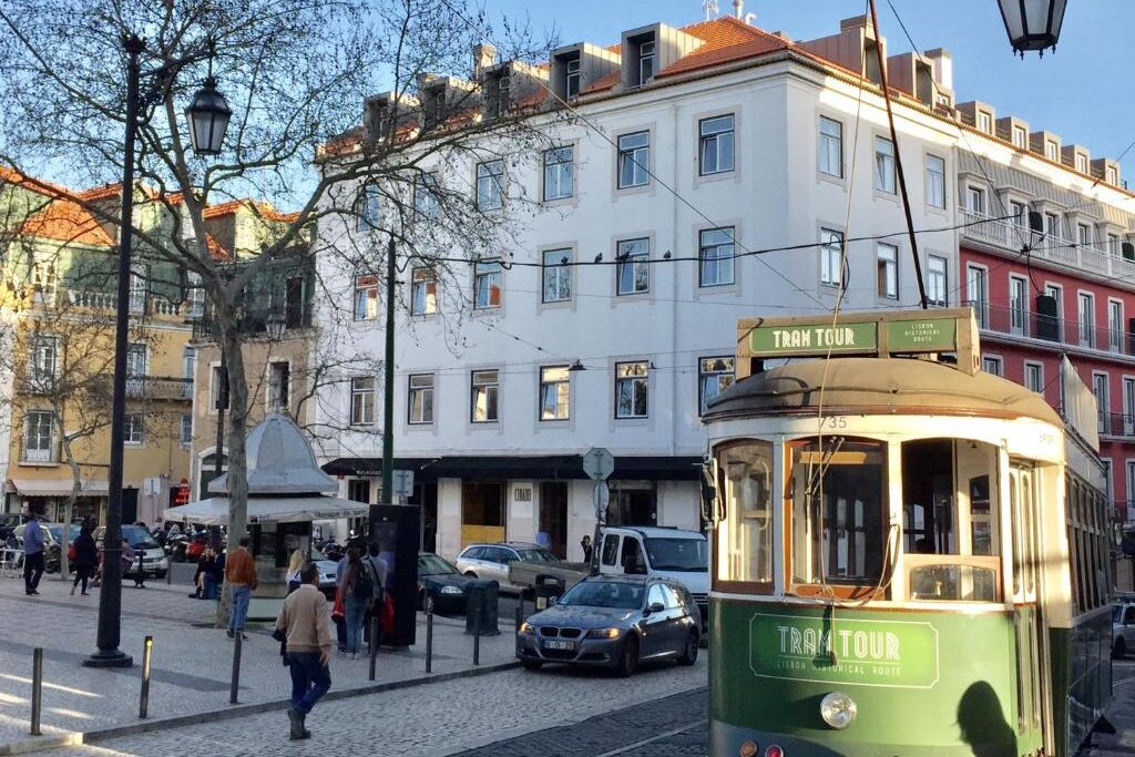 Um elétrico verde passa por uma praça de uma cidade europeia com peões, carros e prédios de vários andares, em frente ao Chiado Arty Flats.