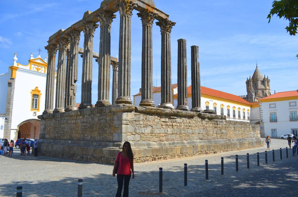 Uma pessoa junto ao Templo Romano de Évora, rodeado por edifícios históricos sob um céu azul.