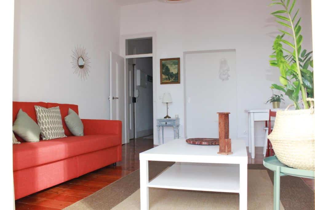 Sala de estar iluminada da Casa da Varanda Simpática com sofá vermelho, almofadas verdes, mesa de centro branca, arte de parede e uma planta. Porta e secretária ao fundo.