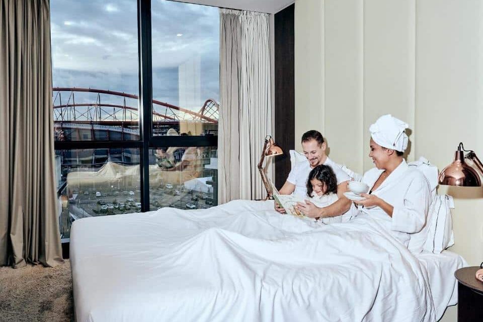 Uma família sentada numa cama de hotel junto a uma grande janela. A mãe segura uma chávena, e o pai e a criança olham para um tablet, no Upon Lisbon Prime Residences.